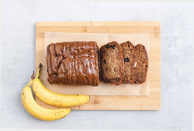 Top down view of Biscoff and chocolate chip banana bread on a wooden board, with three slices cut and two bananas in the bottom left corner