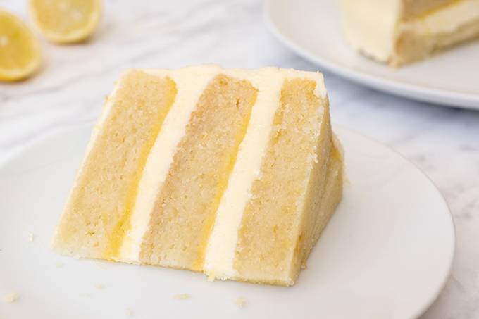 A slice of lemon cake on a plate, with three layers of sponge, lemon curd and lemon buttercream
