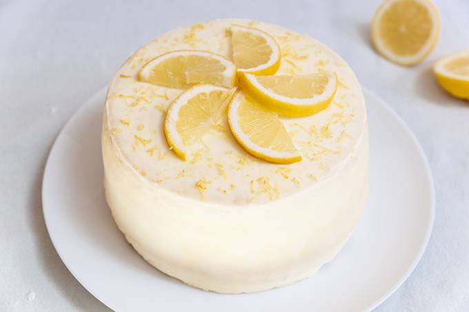 A round lemon layer cake on a white plate, decorated with white icing, lemon slices and zest
