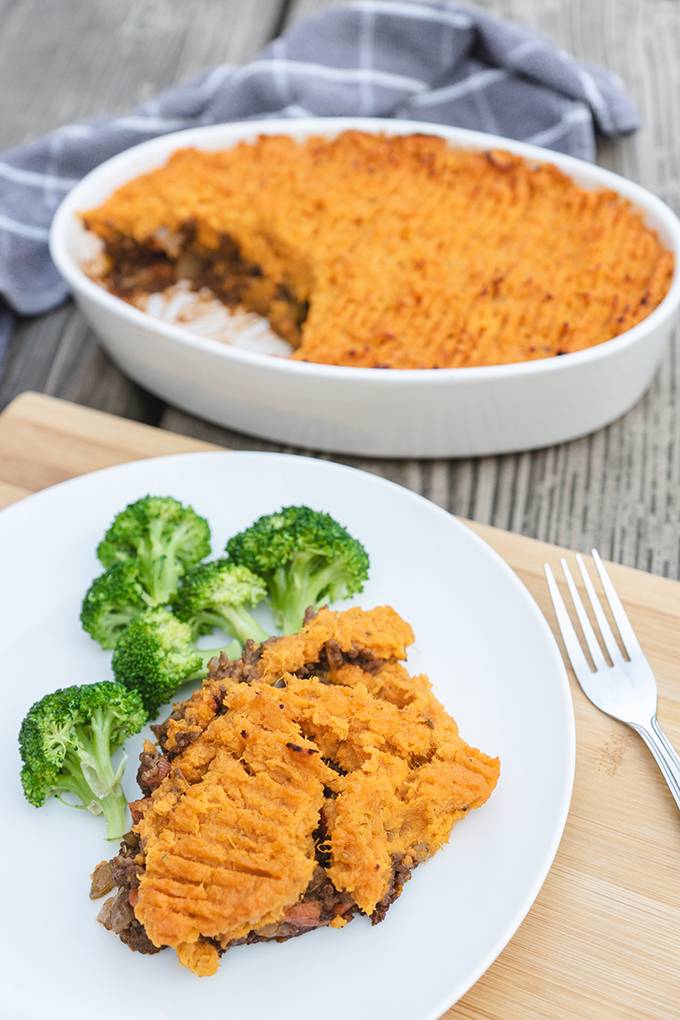 A portion of sweet potato cottage pie on white plate with some broccoli and an oven dish containing the rest of the pie in the background