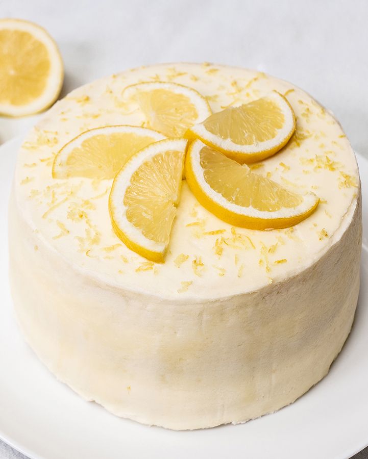 A round lemon layer cake on a white plate, decorated with white icing, lemon slices and zest