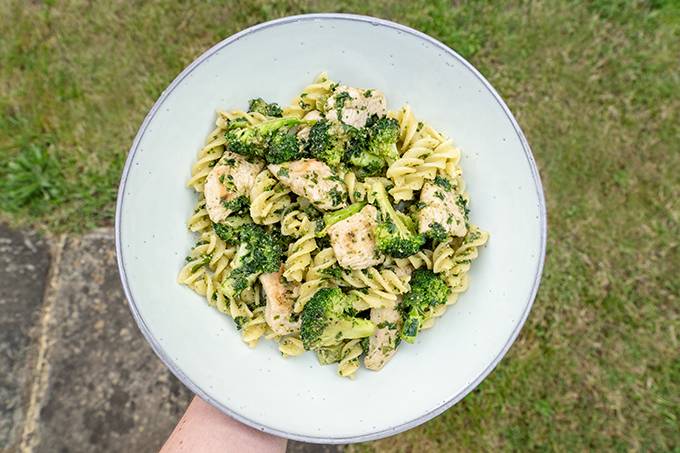A pale blue plate containing pasta, chicken, broccoli and pesto, with green grass in the background.