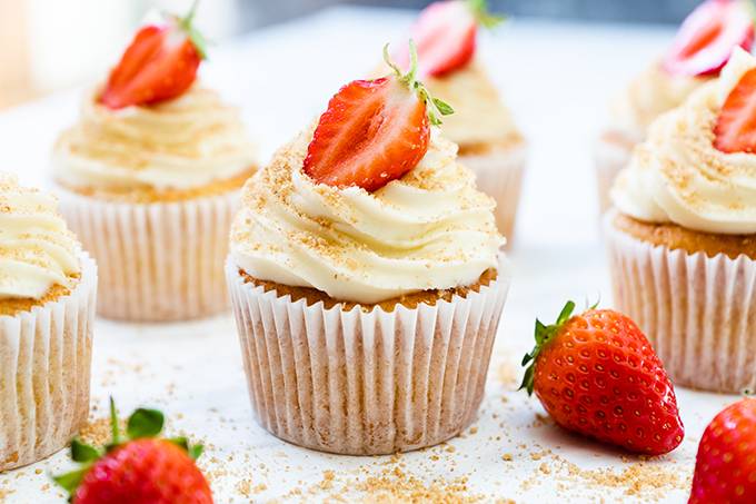 A vanilla cupcake decorated with a white chocolate and cream cheese icing swirl and half a strawberry, surrounded by other cupcakes and strawberries.