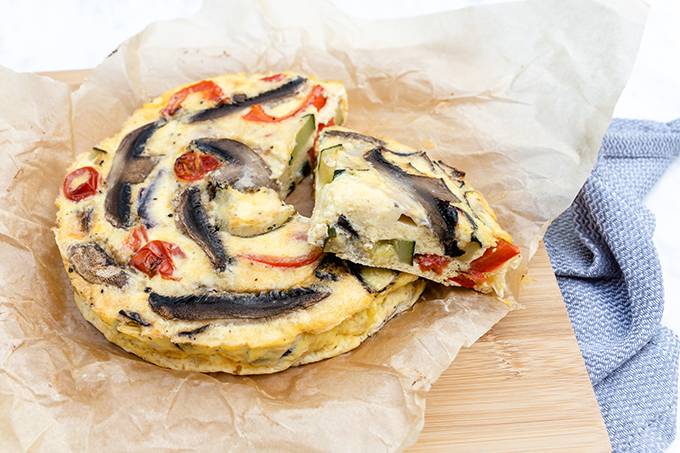 A roasted vegetable frittata on some baking paper on a wooden board, with one slice cut out and resting on top.