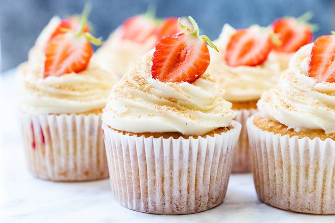 Vanilla cupcakes arranged in rows, each decorated with a white chocolate and cream cheese icing swirl and half a strawberry