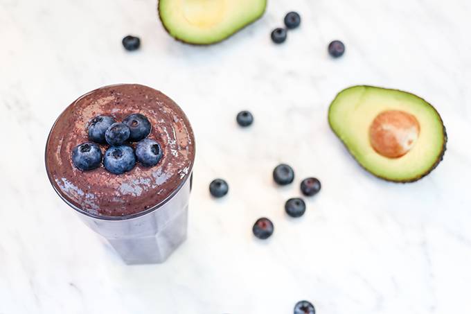 A glass of blueberry avocado smoothie with extra blueberries on top on a white background with a sliced open avocado next to it.