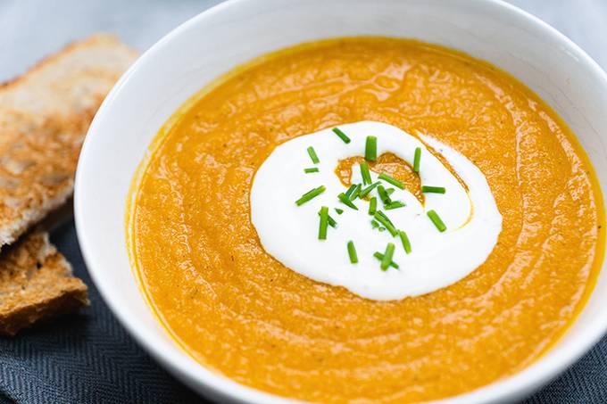 A bowl of smooth orange vegetable soup topped with yoghurt and a sprinkle of chives, with sliced toast next to it.