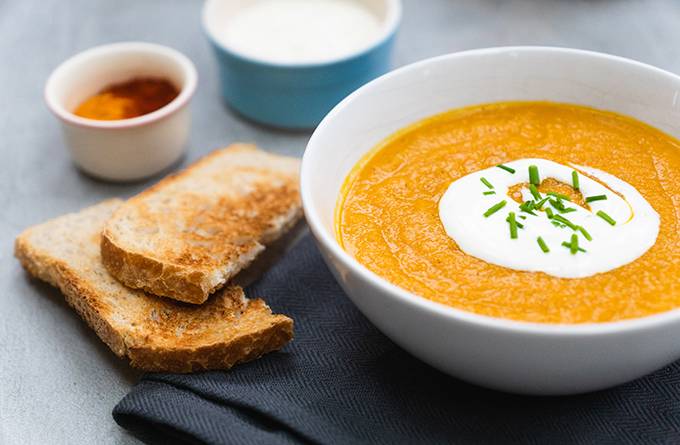 A bowl of smooth orange vegetable soup topped with yoghurt and a sprinkle of chives, with sliced toast next to it.
