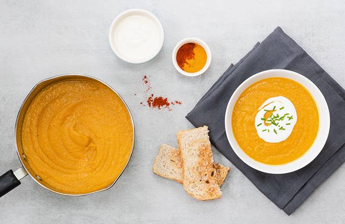 A bowl of smooth orange vegetable soup topped with yoghurt and a sprinkle of chives, with sliced toast and a bigger saucepan of the soup next to it.