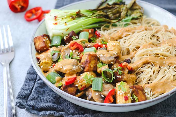 A bowl of noodles with vegetables, tofu and drizzles of peanut sauce on top