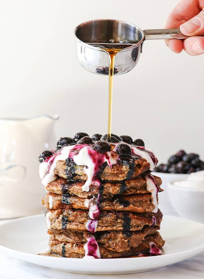 A stack of banana bread pancakes on a white plate, topped with yoghurt and blueberries and being drizzled with maple syrup.