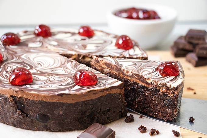 A chocolate biscuit cake with a marbled chocolate topping and cherries on top, with a slice taken out of it.