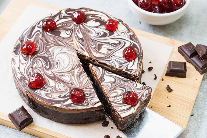 A chocolate biscuit cake with a marbled chocolate topping and cherries on top, with a slice taken out of it.