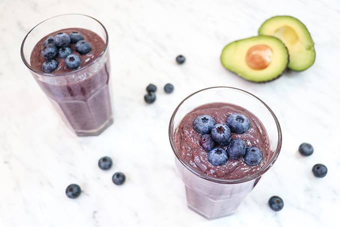 Two glasses of blueberry avocado smoothie with extra blueberries on top on a white background with a sliced open avocado next to it.