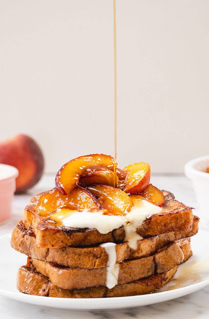 A plate of French toast topped with caramelised peaches and yoghurt, and maple syrup being poured in top