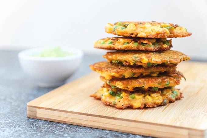 A stack of fried vegetable fritters on a wooden board, next to a white bowl of guacamole