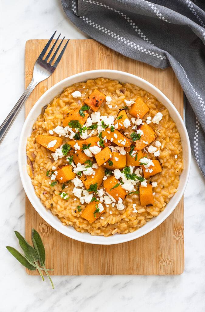 Top down view of a white bowl on a wooden board, containing roasted butternut squash risotto which is topped with crumbled feta cheese and fresh sage.