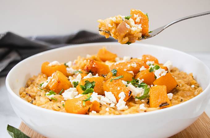 A white bowl containing roasted butternut squash risotto topped with crumbled feta cheese and fresh sage, with a forkful held above the bowl.