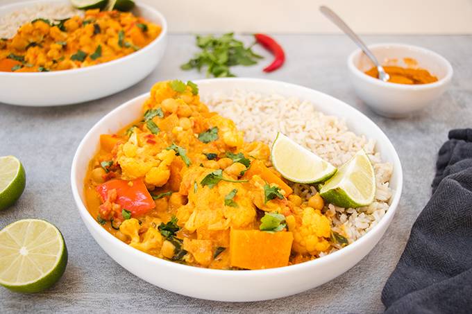 A white bowl containing vegetable curry, rice and two wedges of lime