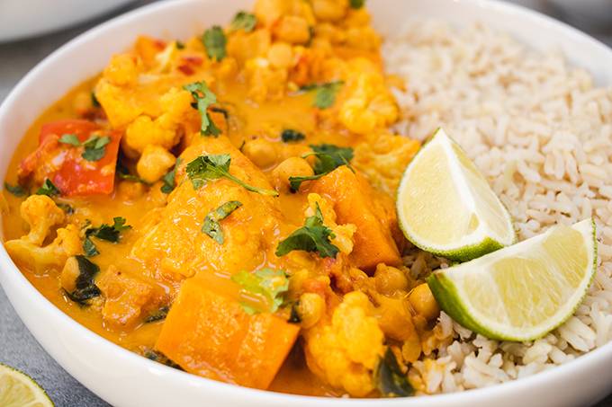 Close up of a bowl of vegetable curry with rice and two wedges of lime