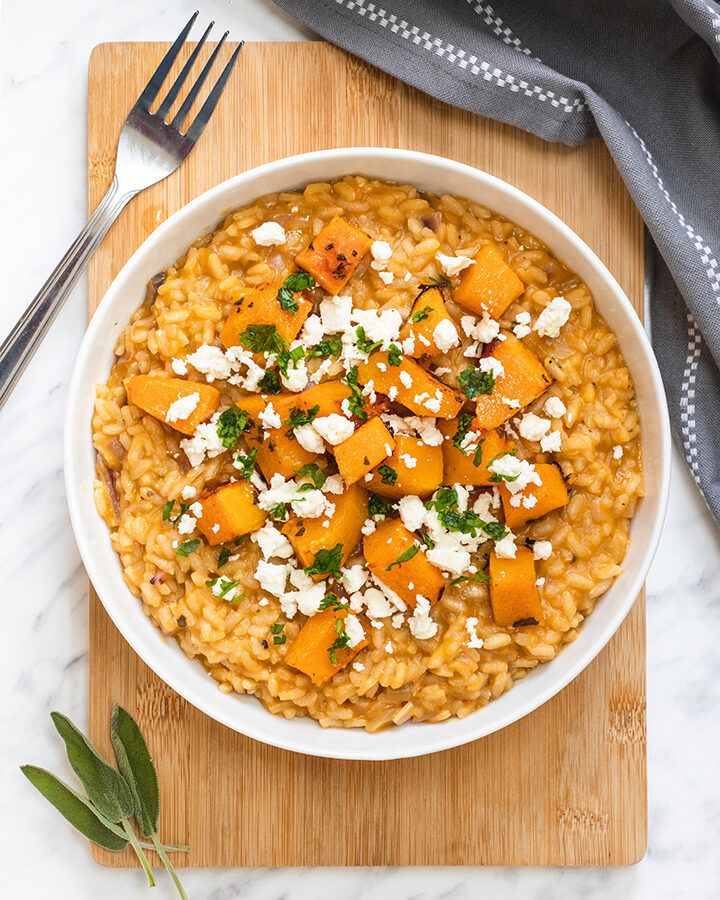 Top down view of a white bowl on a wooden board, containing roasted butternut squash risotto which is topped with crumbled feta cheese and fresh sage.