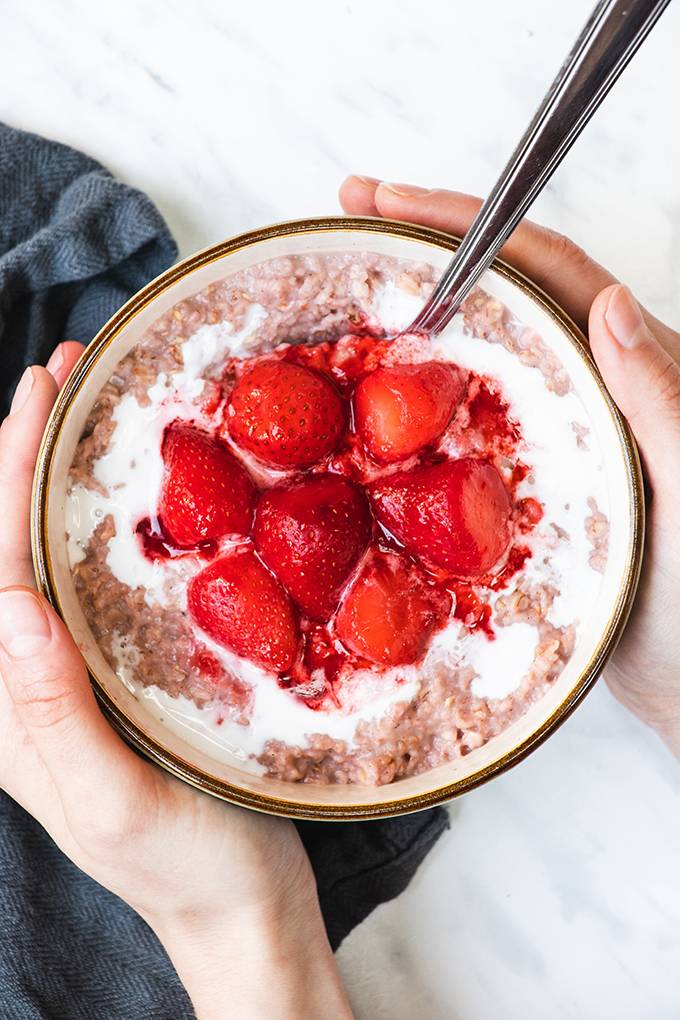 Top down view of a bowl of porridge topped with coconut cream and cooked strawberries, being held in a person's hands