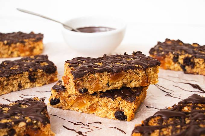 Two chocolate apricot flapjacks stacked, surrounded by other flapjacks and with a pot of melted dark chocolate behind them.