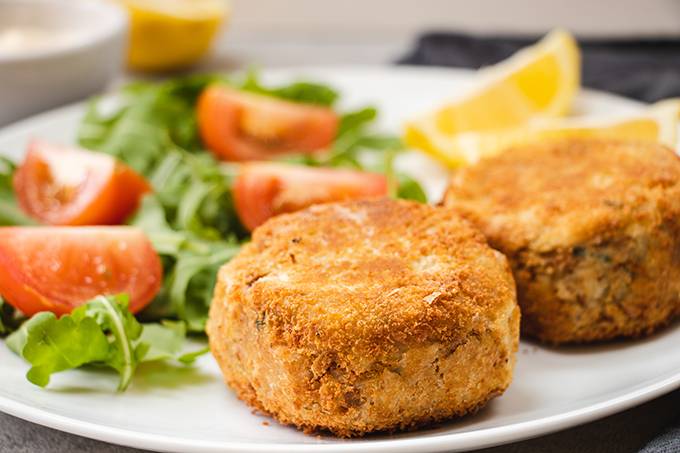 Two tuna fishcakes on a white plate with some salad and lemon wedges in the background