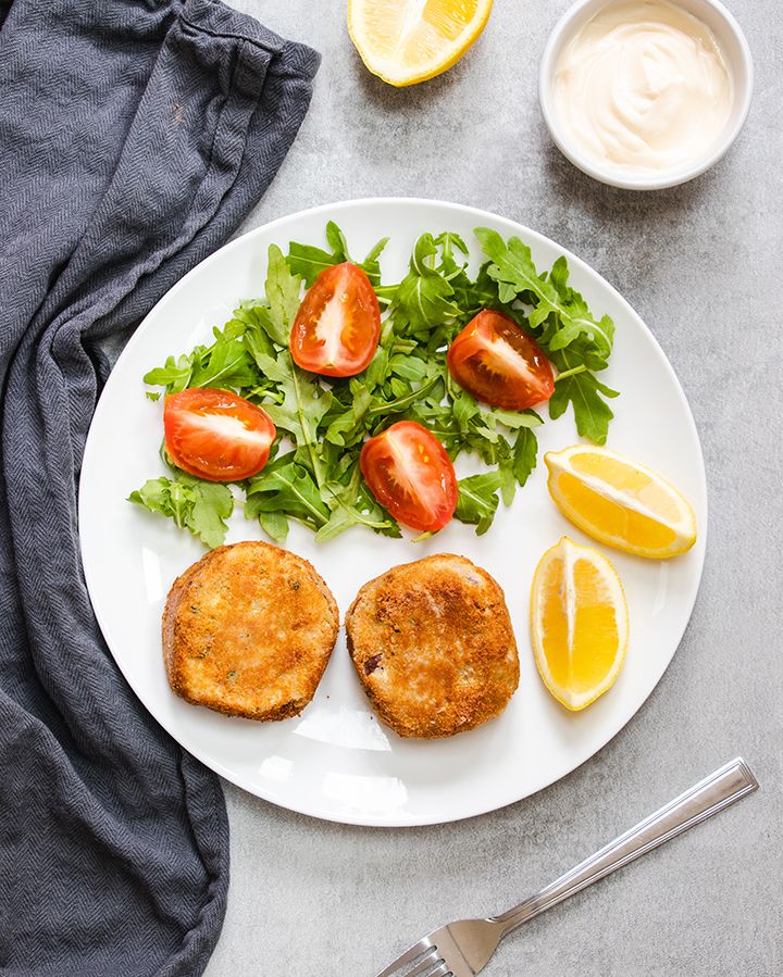 Top down view of a white plate containing two tuna fishcakes, salad and lemon wedges