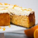An orange and almond cake on a white cake stand with a slice being taken out of it
