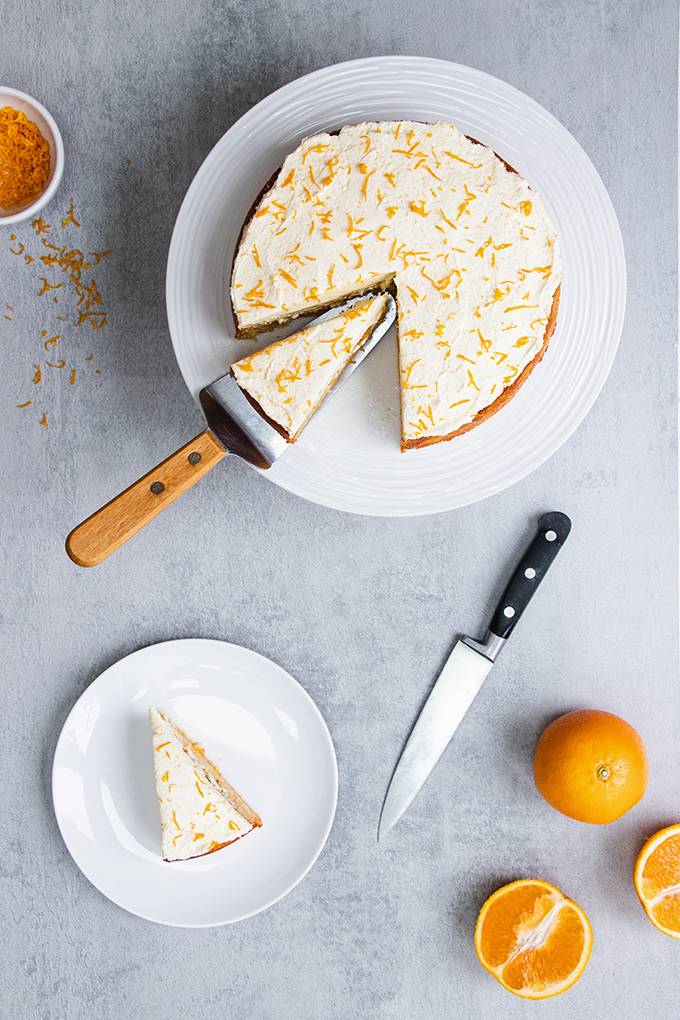 Top down view of an orange and almond cake with icing and orange zest on top, with one slice put on a separate plate