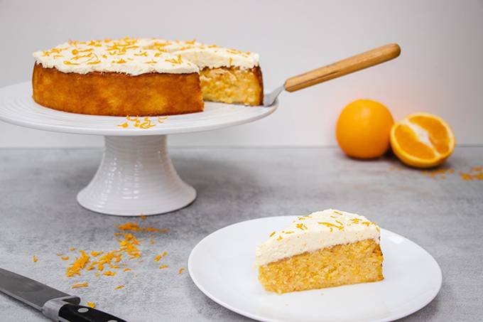 An orange and almond cake on a white cake stand, with one slice separately on a small white plate