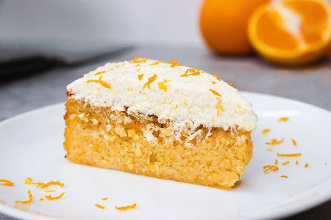 Close up of a slice of orange and almond cake on a white plate, with white buttercream icing and orange zest on top