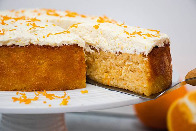 An orange and almond cake on a white cake stand with a slice being taken out of it