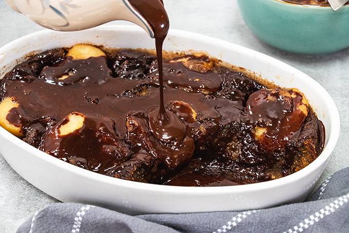 A pear and ginger pudding in a white dish, with chocolate sauce being poured on top from a jug.