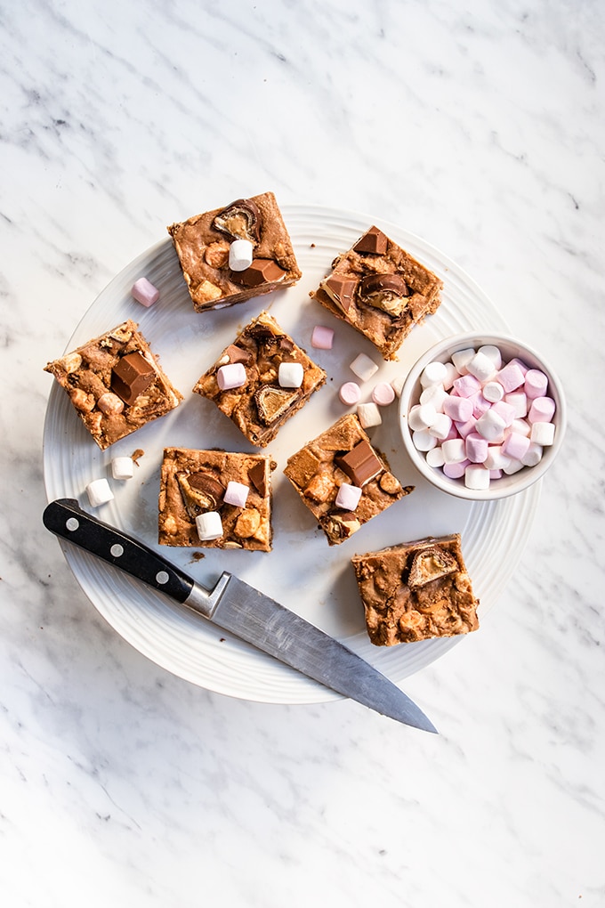 Top down view of seven Kinder rocky road squares on a white cake stand, with a knife and small bowl of mini marshmallows next to them.