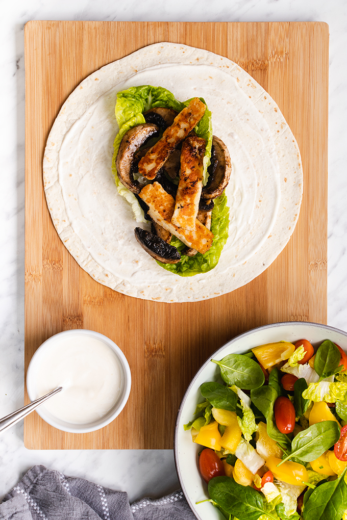 Top down view of an open tortilla wrap with mushroom and halloumi, laid out on a wooden board, next to a large bowl of salad and a small pot of yoghurt mayo.