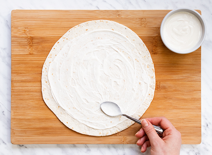 Top down view of a tortilla wrap on a wooden board, being spread with yoghurt mayo.