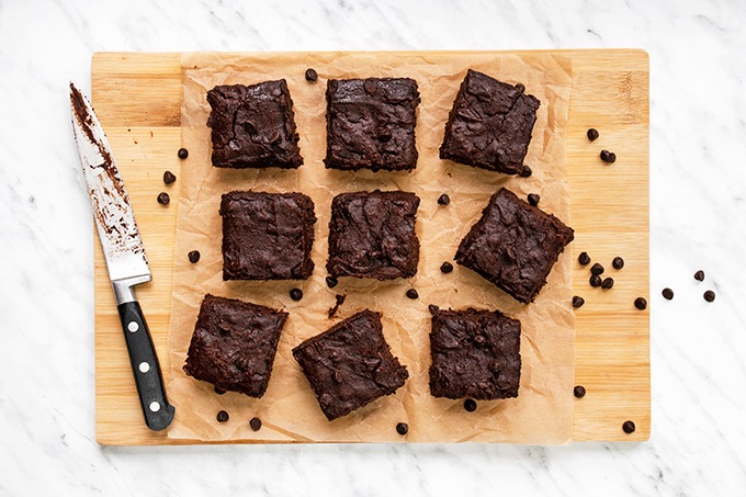 Top down view of fudgy vegan brownies on a wooden board, with a knife next to them and chocolate chips scattered around.