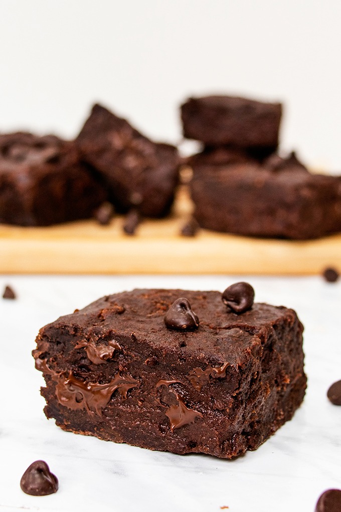 Close up of a gooey vegan date brownie, with chocolate chips scattered around and other brownies on a wooden board in the background.