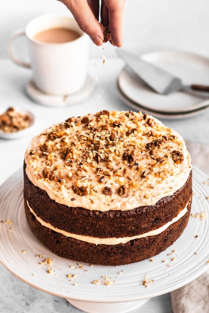 A carrot cake with frosting, being sprinkled with chopped walnuts.